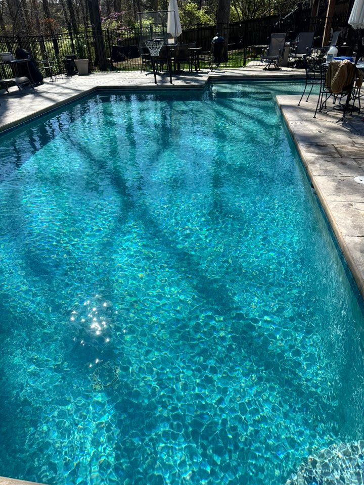 A rectangular, turquoise pool with sun reflections. Tables and chairs are near the pool on a stone patio.
