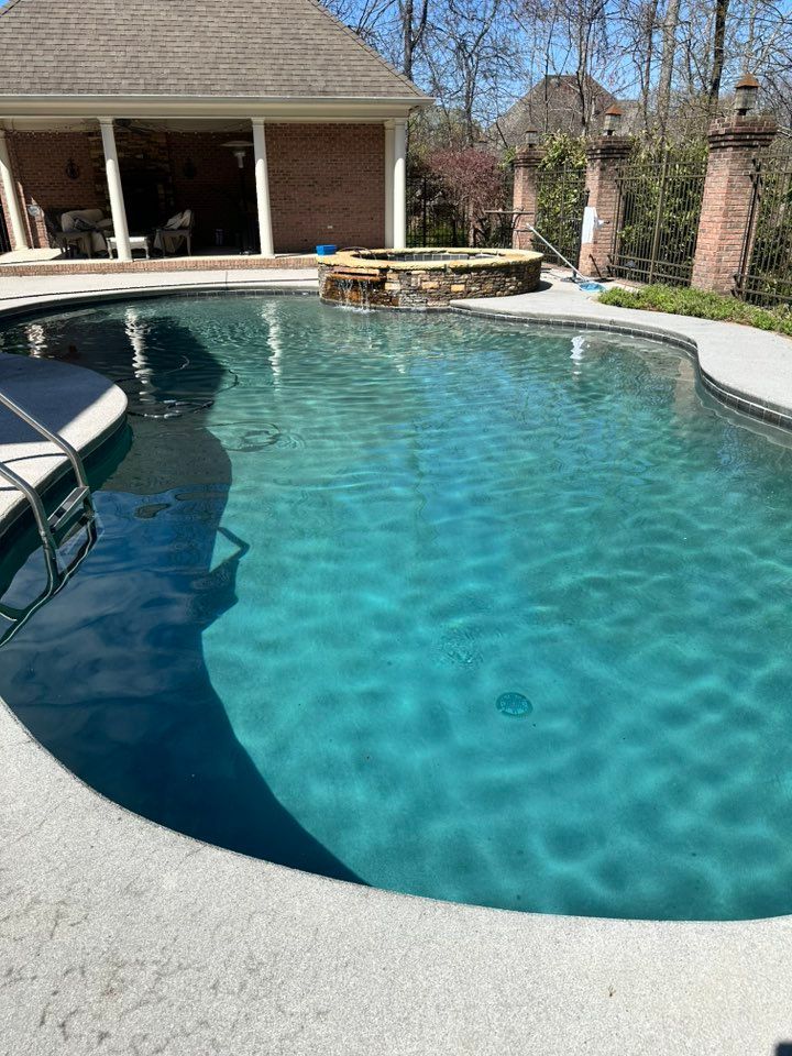 A turquoise swimming pool with a stone feature, surrounded by a concrete patio, next to a brick building.