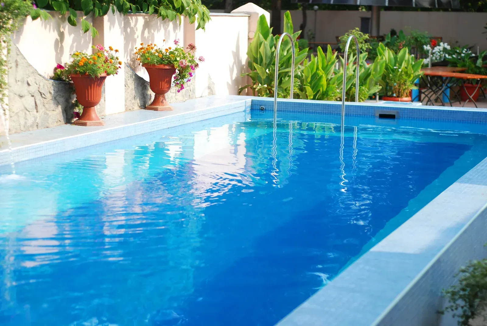 Blue-tiled swimming pool with clear water, surrounded by plants and two flower pots.