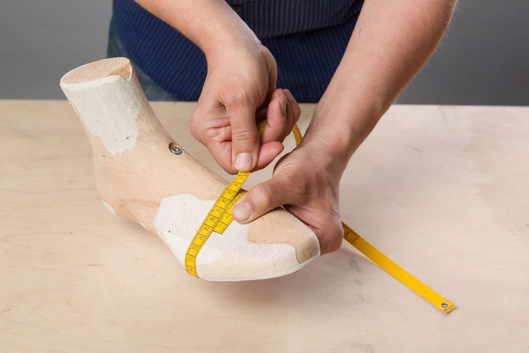 Un paio di mani utilizzano un metro a nastro giallo per verificare la larghezza del piede di un manichino di colore chiaro su una superficie di legno.