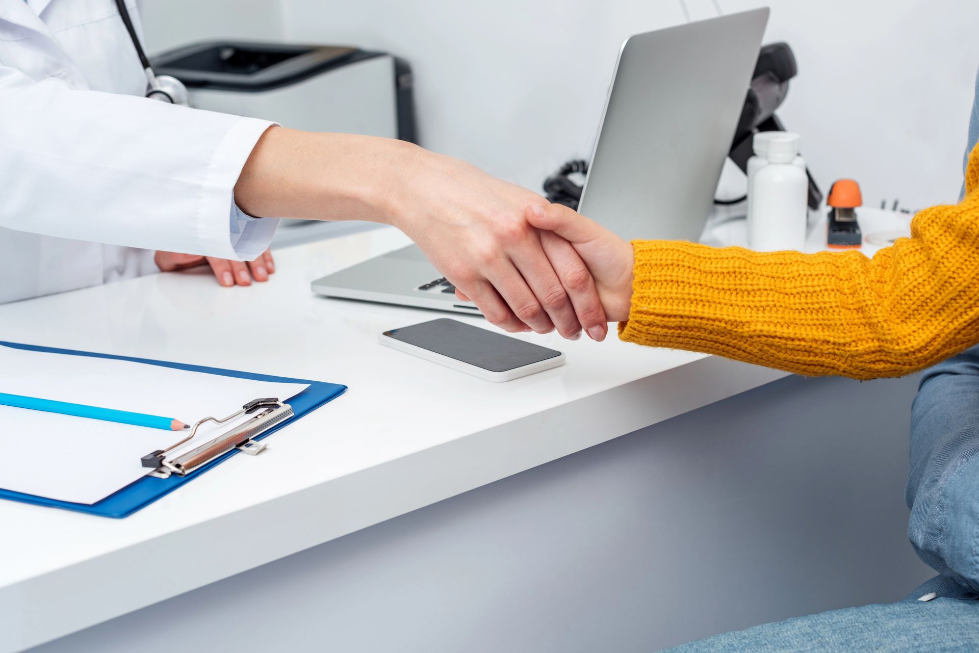 In uno studio medico, un medico in camice bianco stringe la mano a un paziente con un maglione giallo, seduti attorno a una scrivania.