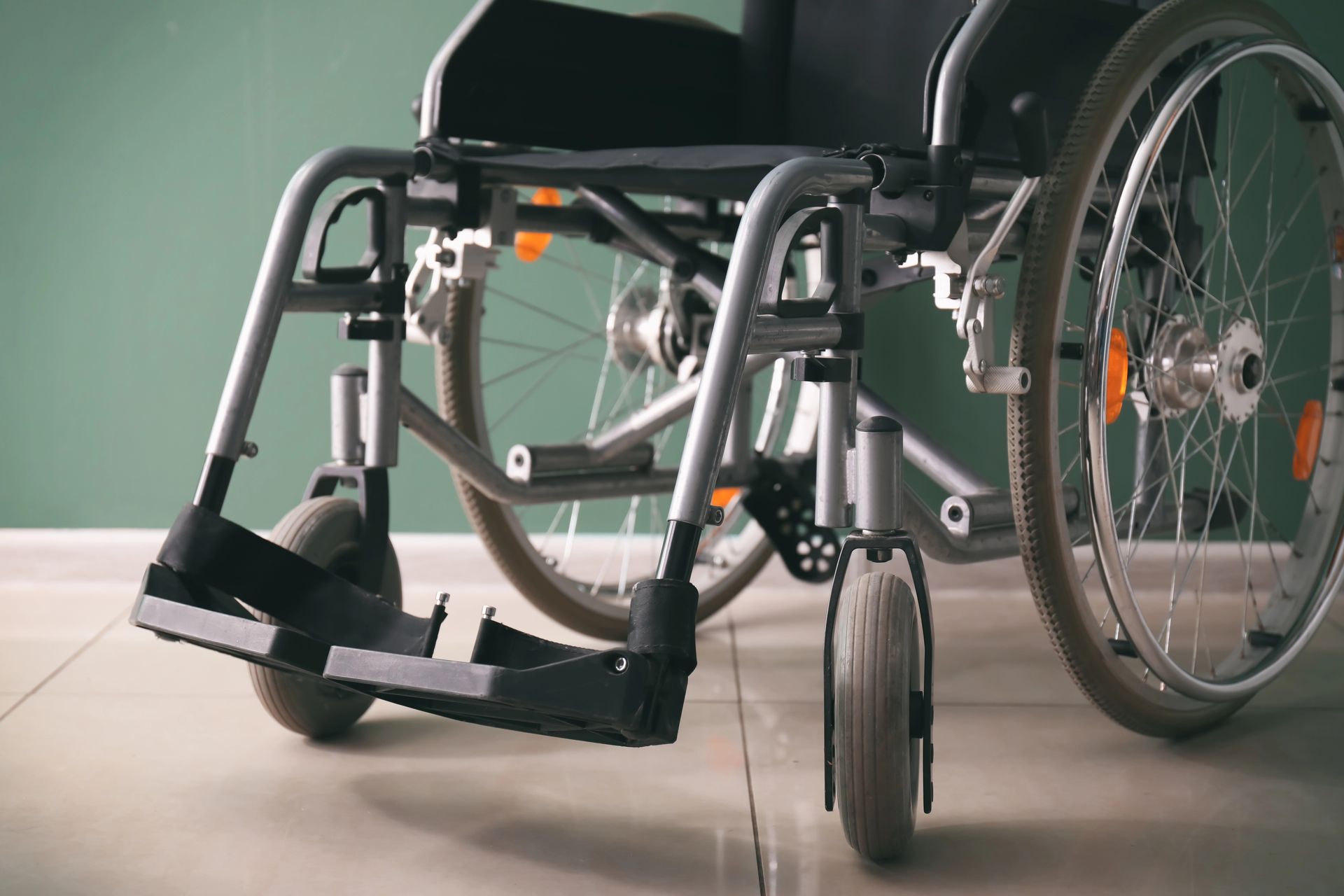 Una sedia a rotelle manuale grigia con poggiapiedi nero, posizionata su un pavimento piastrellato contro una parete verde salvia.