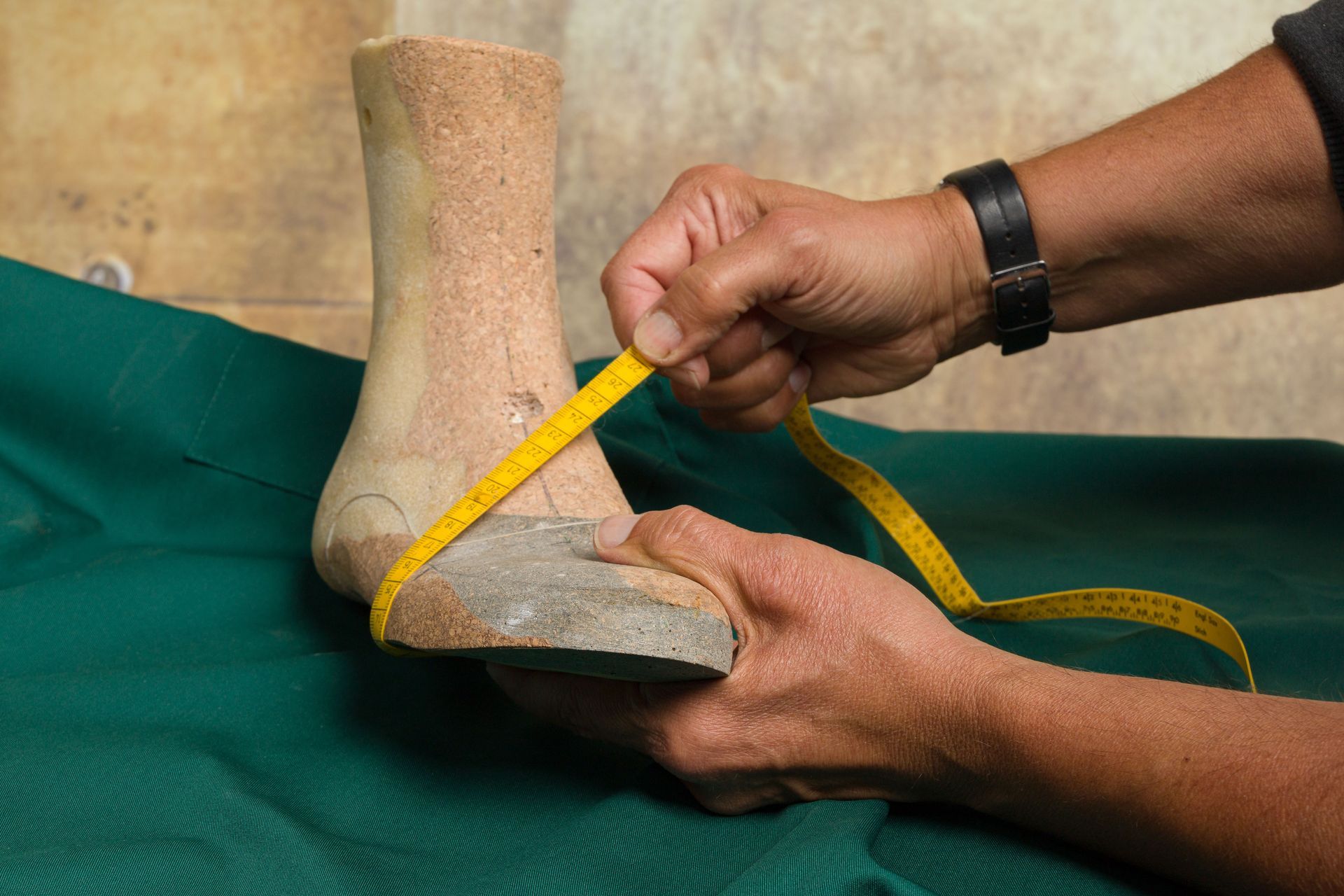 Delle mani misurano una forma per scarpe beige con un metro a nastro giallo su uno sfondo di stoffa verde.