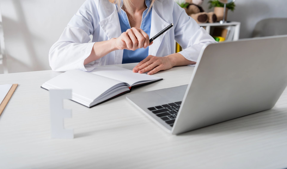 Un professionista in camice bianco è seduto a una scrivania con un computer portatile, un quaderno aperto e una lettera