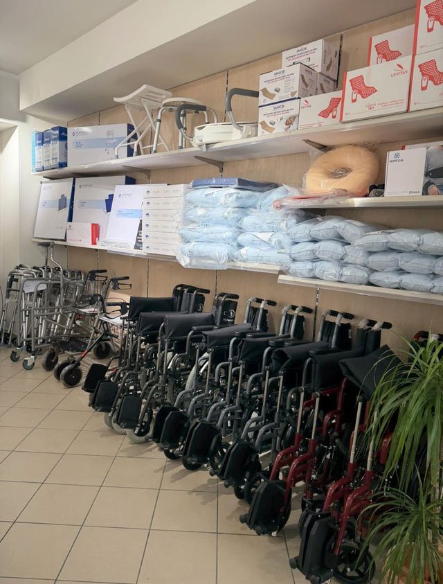 Un negozio di forniture mediche con sedie a rotelle e deambulatori sul pavimento e vari ausili per la mobilità sugli scaffali.
