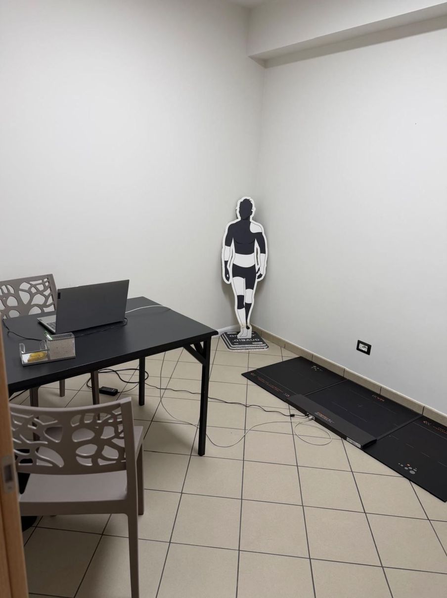 Una stanza clinica con un tavolo nero su cui è appoggiato un computer portatile, delle sedie e un bersaglio anatomico a forma umana posizionato contro una parete.