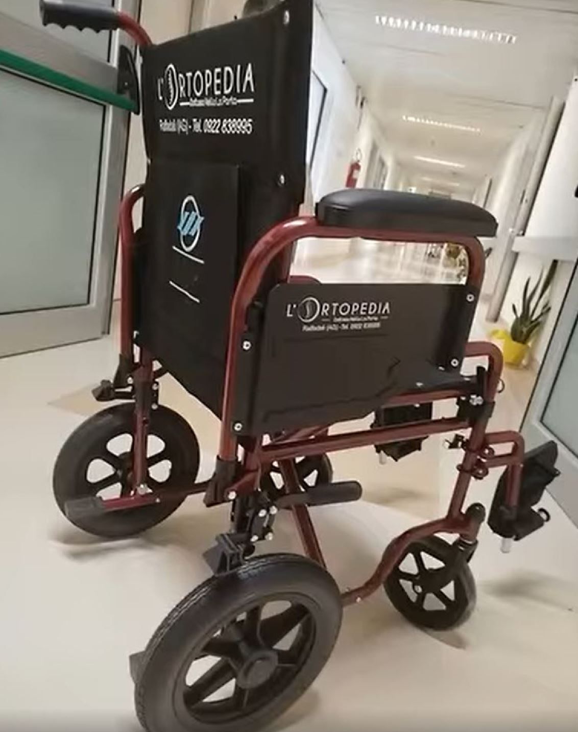 Una sedia a rotelle pieghevole color bordeaux con logo nero, posizionata in un luminoso corridoio d'ospedale.