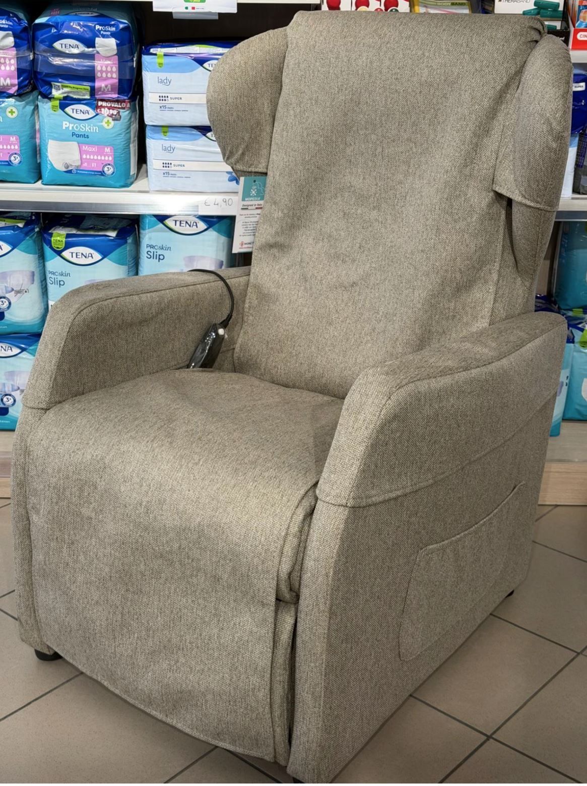 Una poltrona reclinabile beige con meccanismo di sollevamento elettrico e alette sul poggiatesta, posizionata nel corridoio di un negozio con scaffali di prodotti alle spalle.