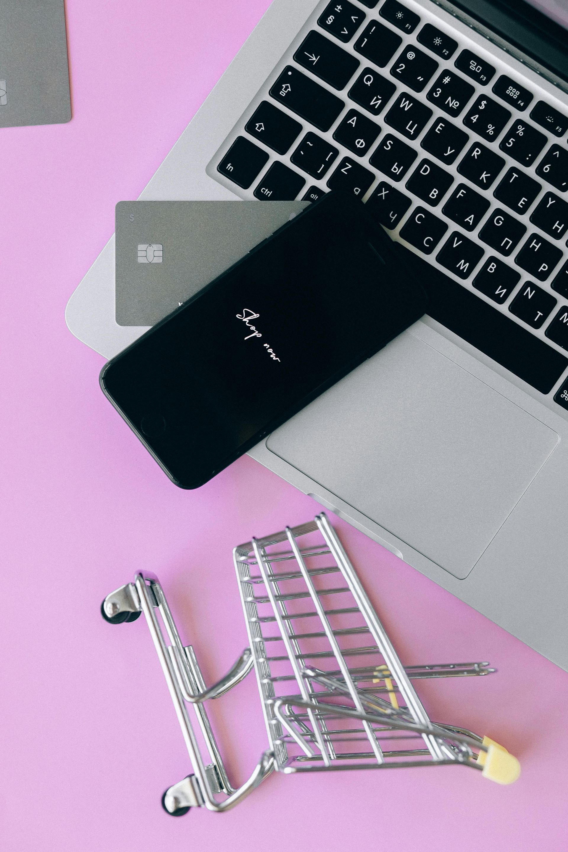 Laptop, telefone, cartão de crédito e carrinho de compras em miniatura em uma superfície rosa, sugerindo compras online.