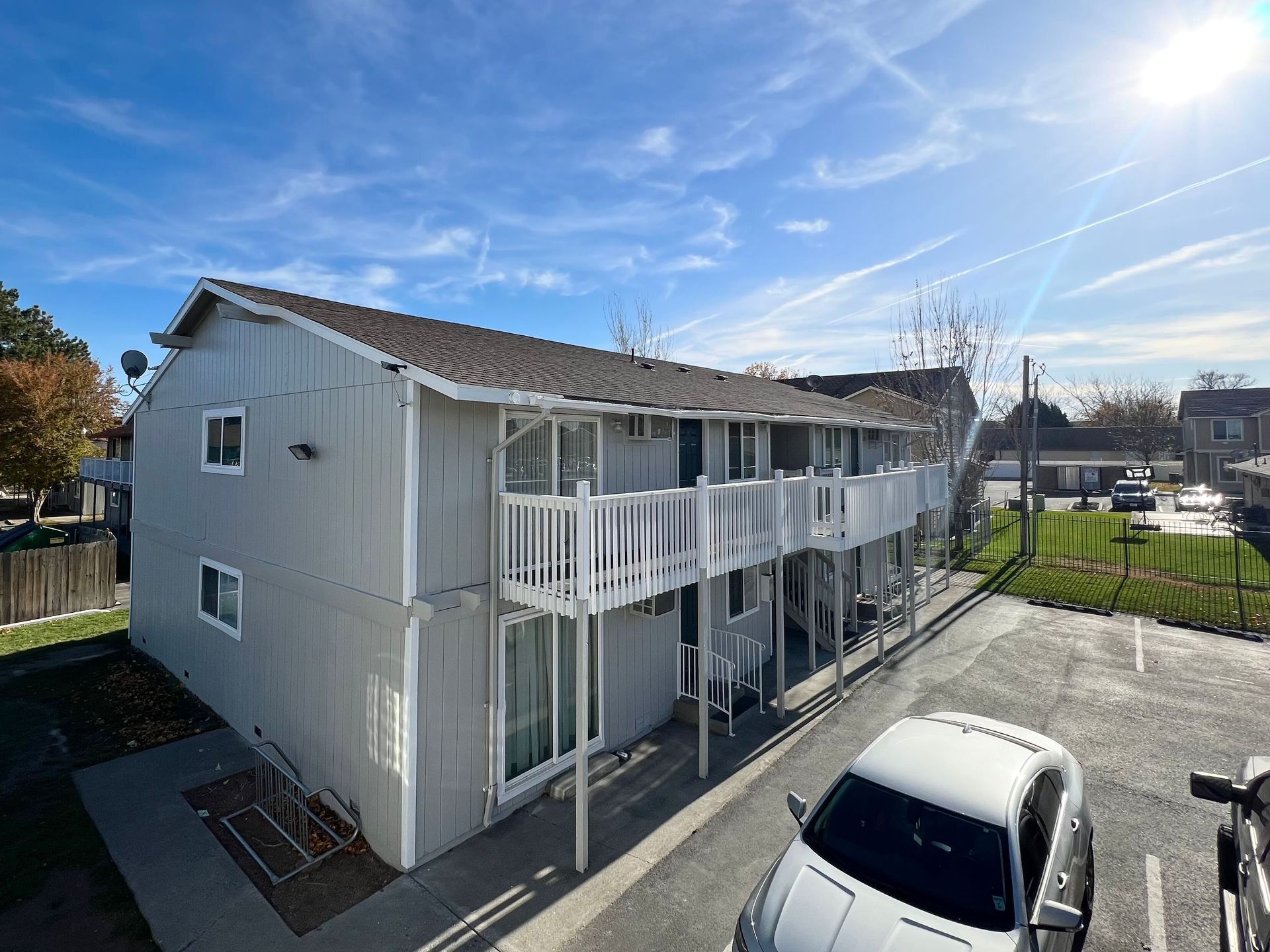 Sunlit exterior of a white two-story apartment building with balconies and parked cars in a lot