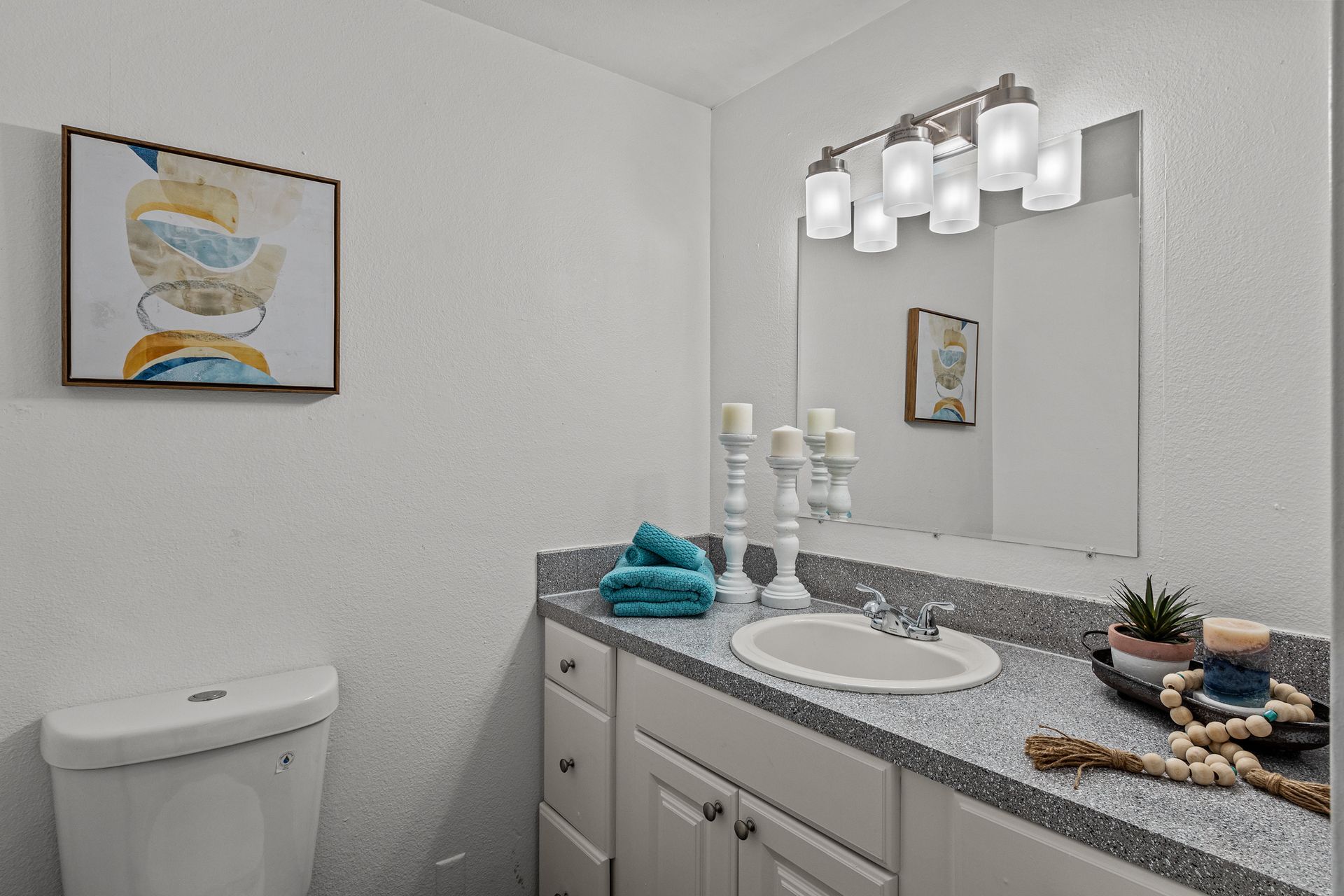 Small bathroom vanity with granite countertop, white cabinets, mirror, sconce lights, and wall art.