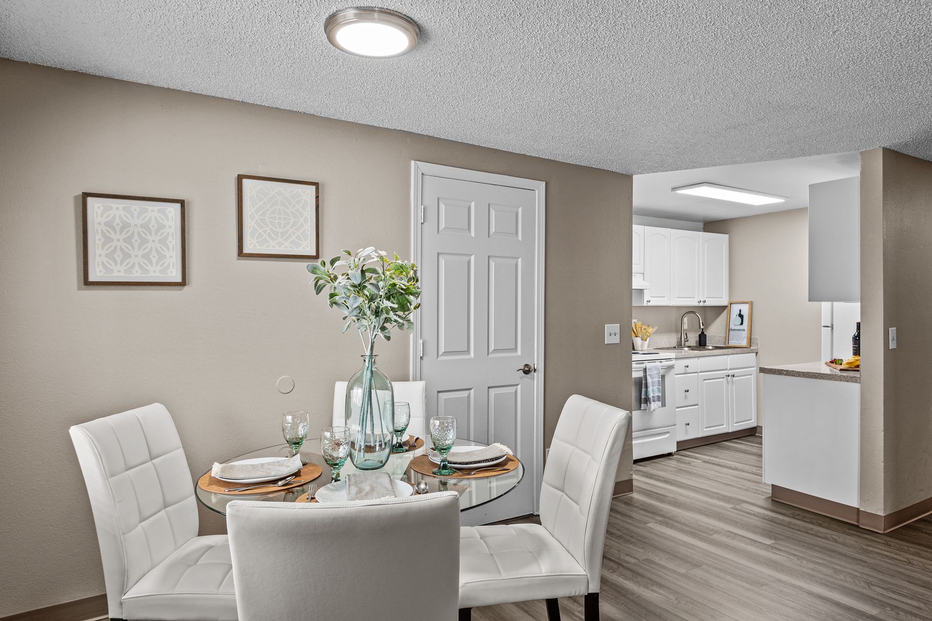 Dining area with round table set for a meal, beige chairs, wall art, and view into a bright kitchen.