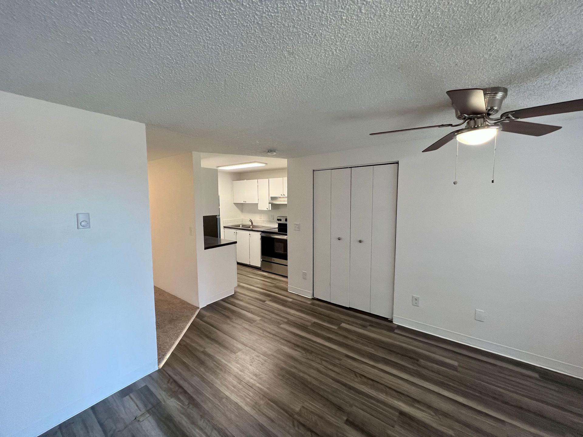 Empty living room with wood-look floors, ceiling fan, and view into a bright kitchen.