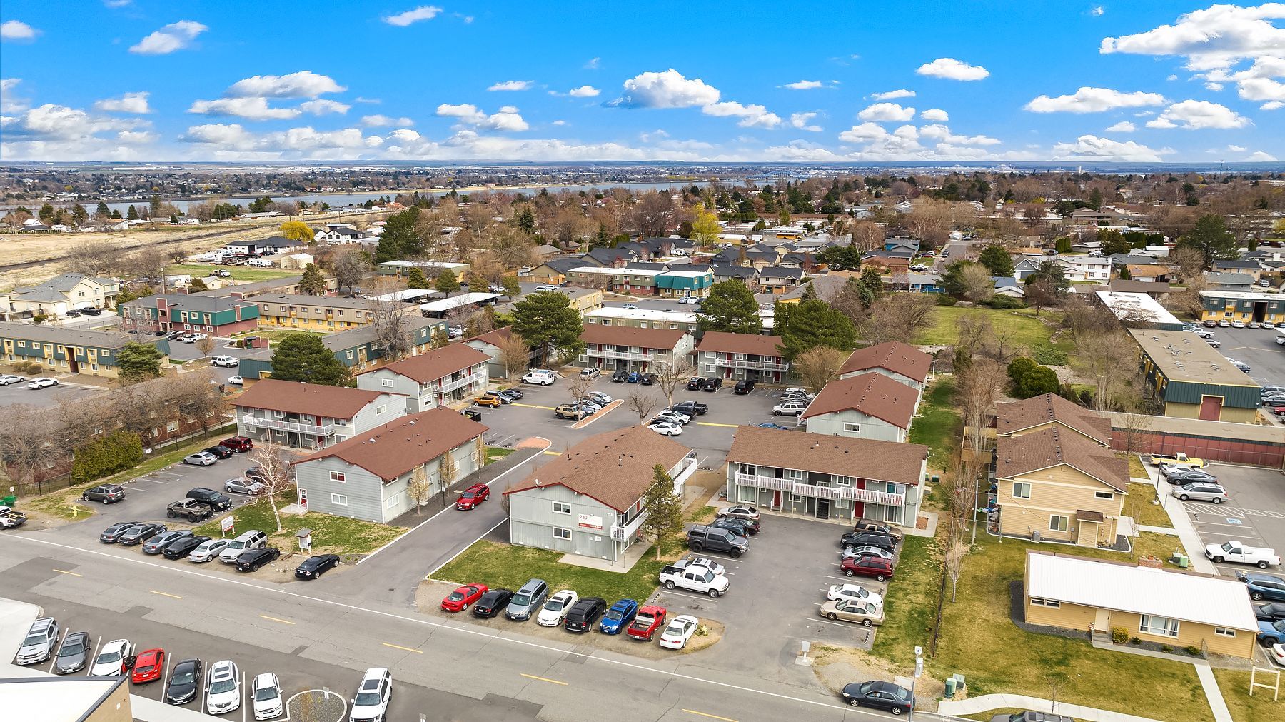 Aerial view of a suburban apartment complex with parked cars, roads, and surrounding houses under a clear sky