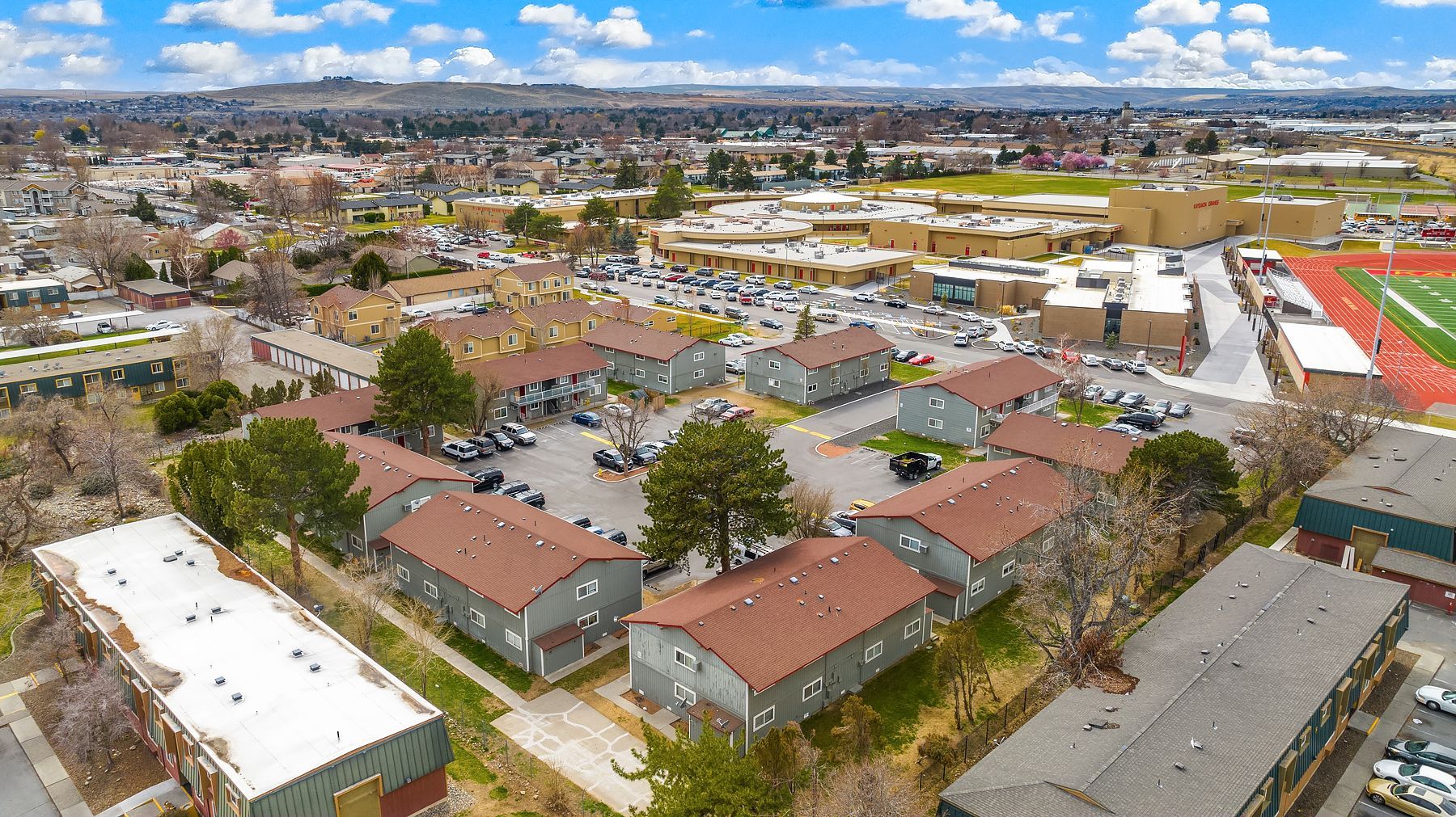 Aerial view of a suburban neighborhood with houses, streets, parking lots, and a distant city skyline under a blue sky.