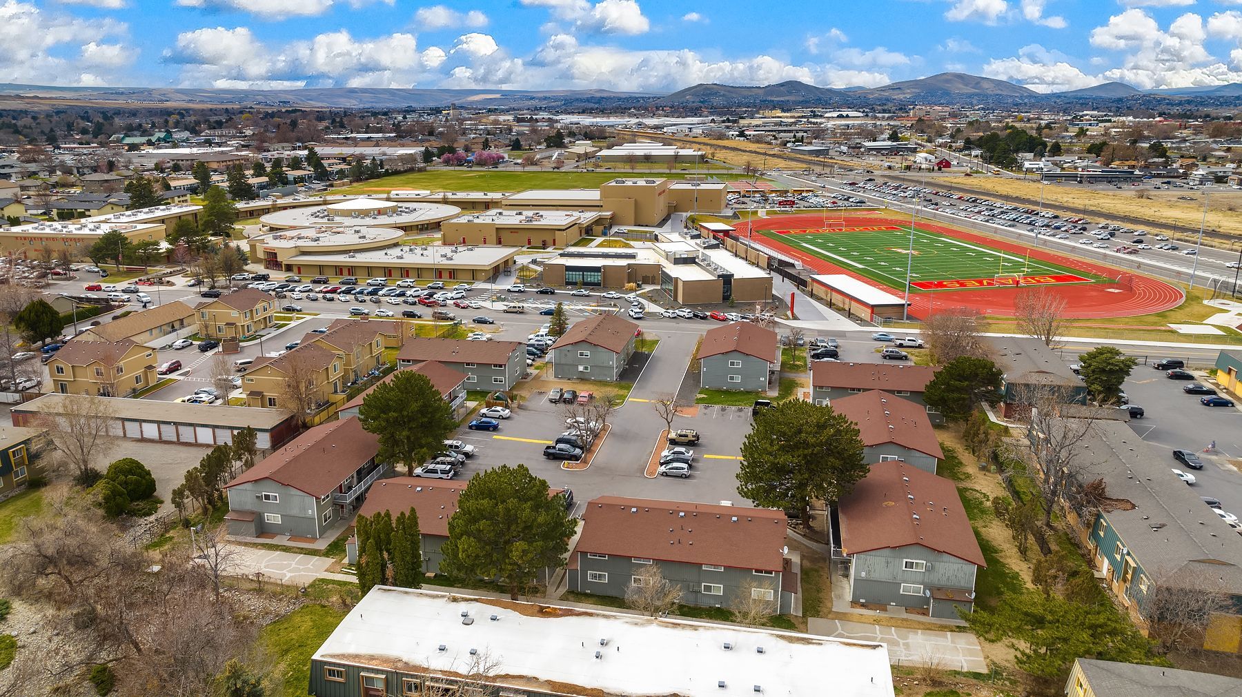 Aerial view of a school campus with buildings, parking lots, and a football field under a cloudy sky.