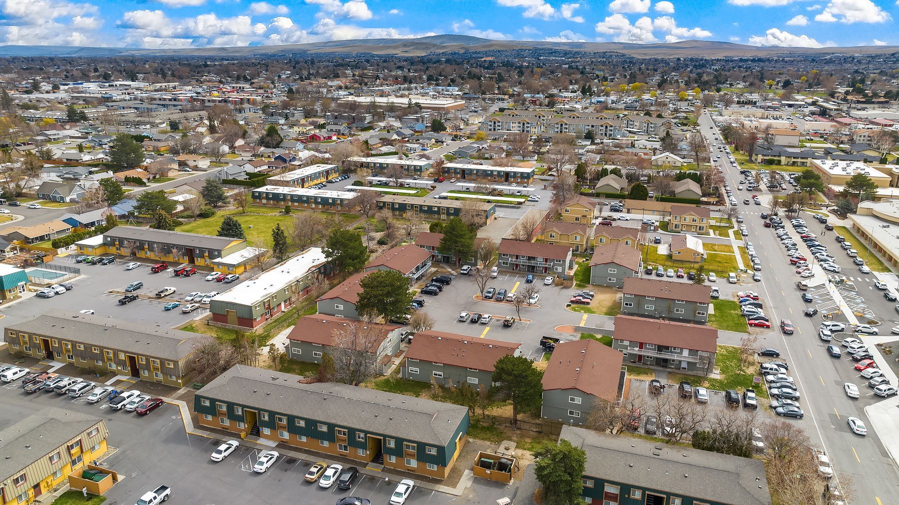 Aerial view of a suburban neighborhood with homes, streets, parked cars, and a distant city skyline under blue sky.