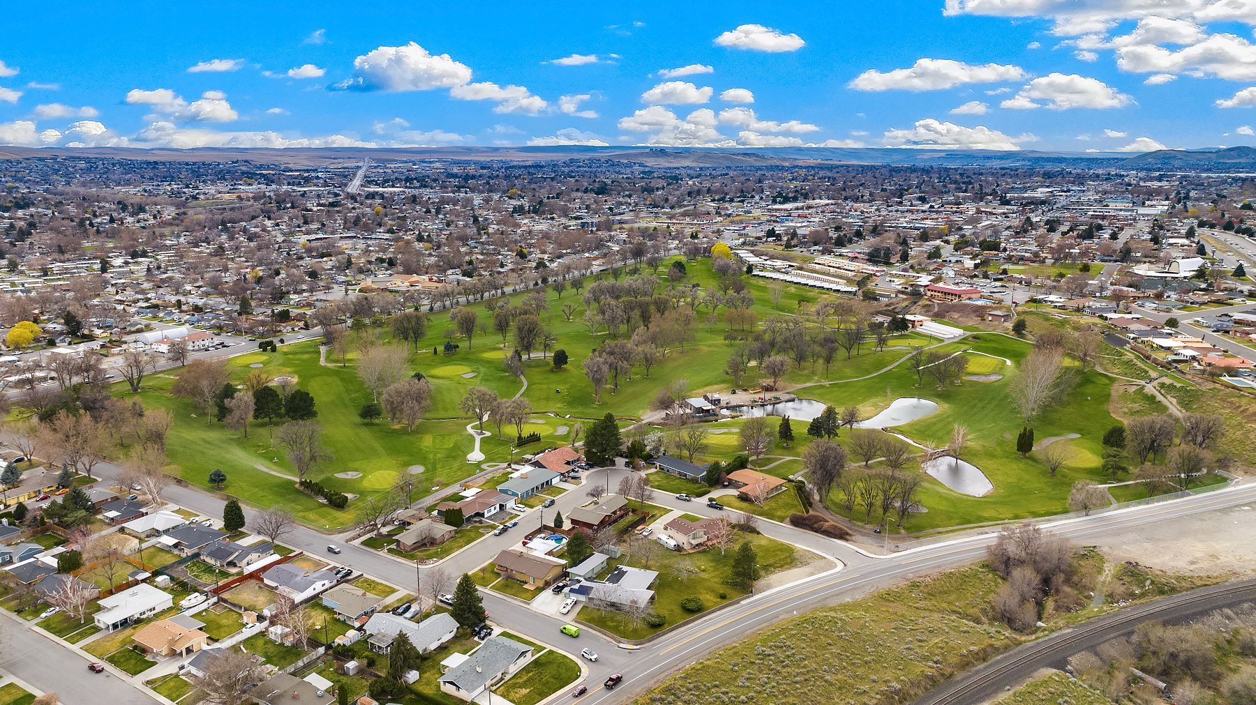 Aerial view of a green park and suburban neighborhood under a blue sky with scattered clouds.
