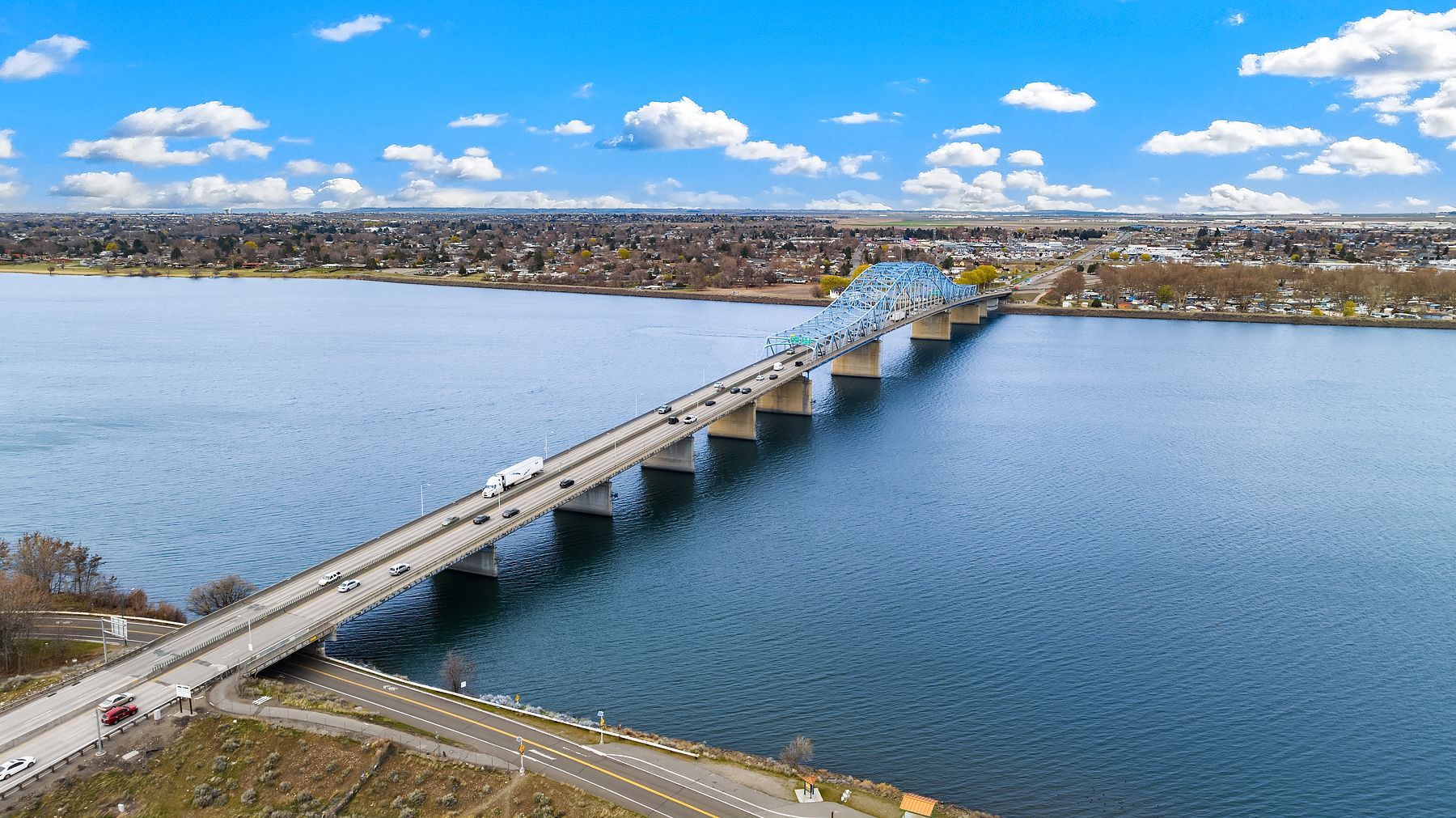A long bridge spans a wide blue river toward a distant shoreline under a partly cloudy sky.