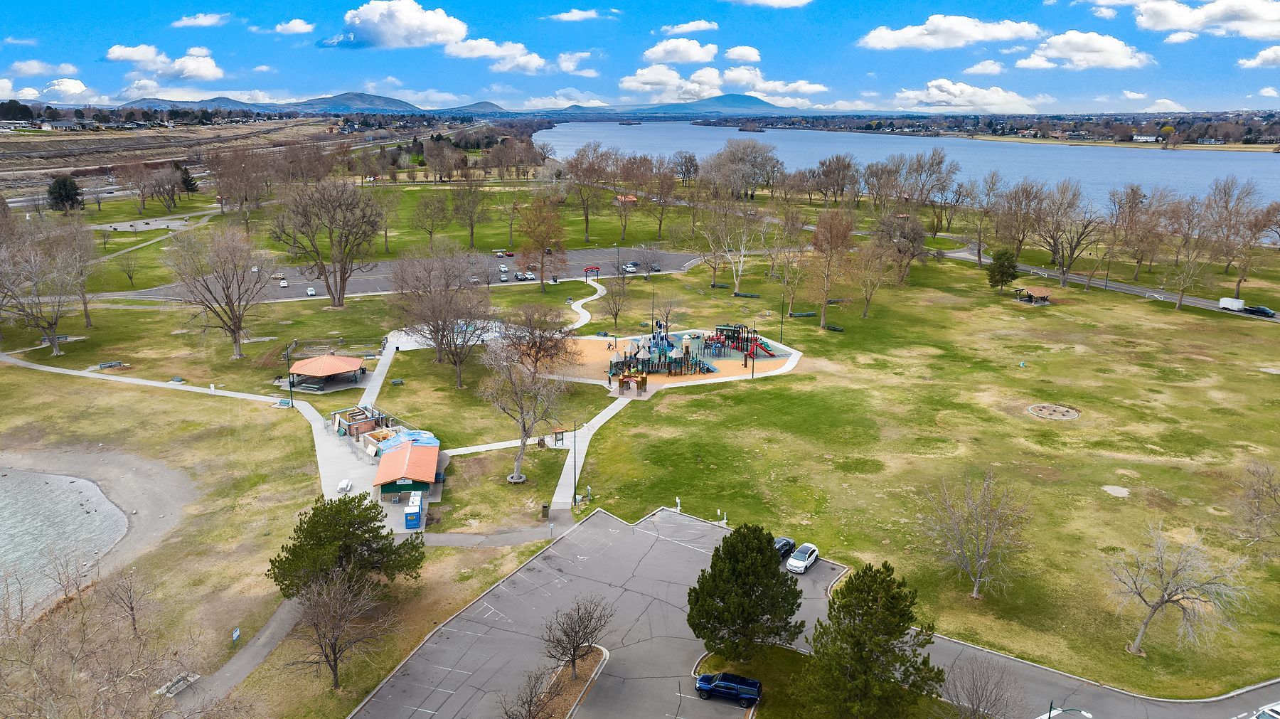 Aerial view of a park by a river, with trees, paths, and small buildings under a cloudy sky.