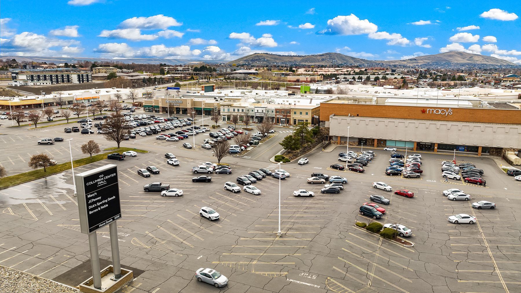 Aerial view of a crowded parking lot beside a large shopping center under a blue sky.