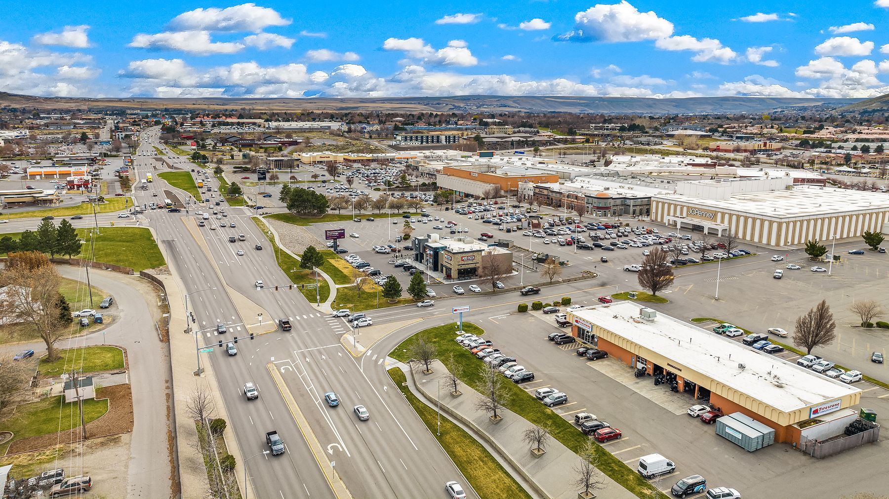 Aerial view of a busy commercial district with roads, parking lots, and shopping centers under a cloudy blue sky