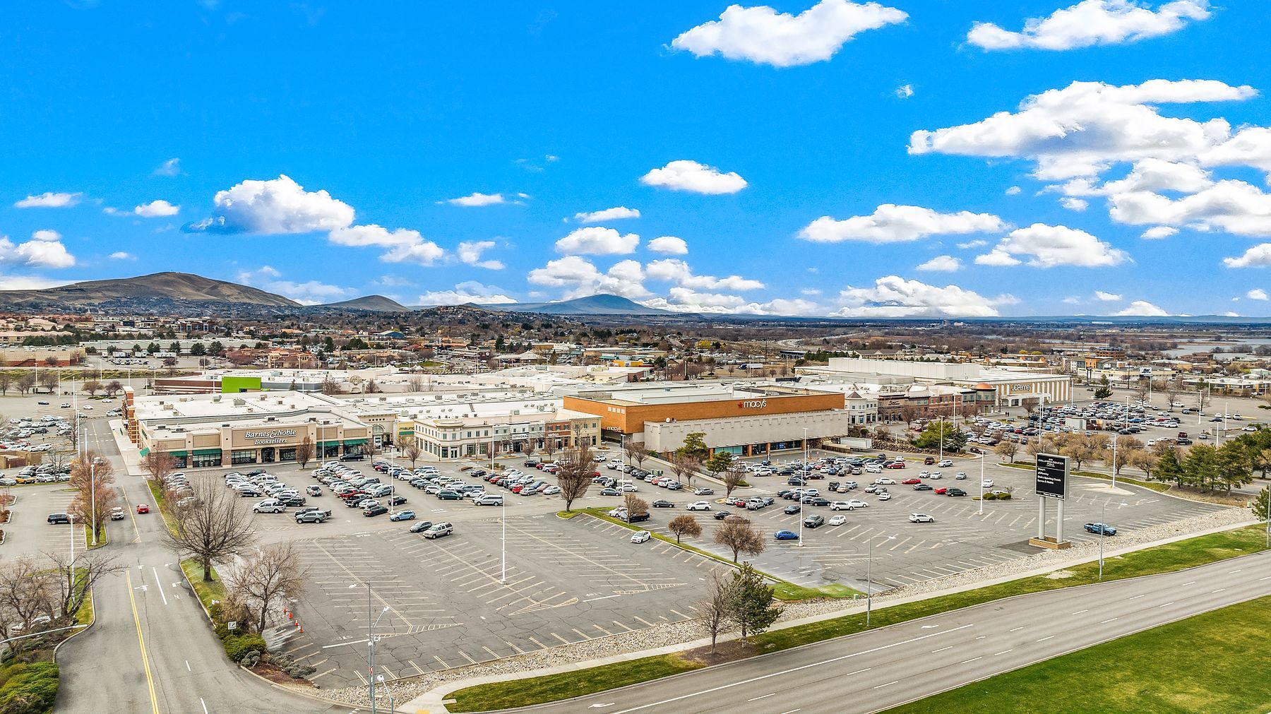 Aerial view of a large shopping center parking lot under a bright blue sky with scattered clouds.