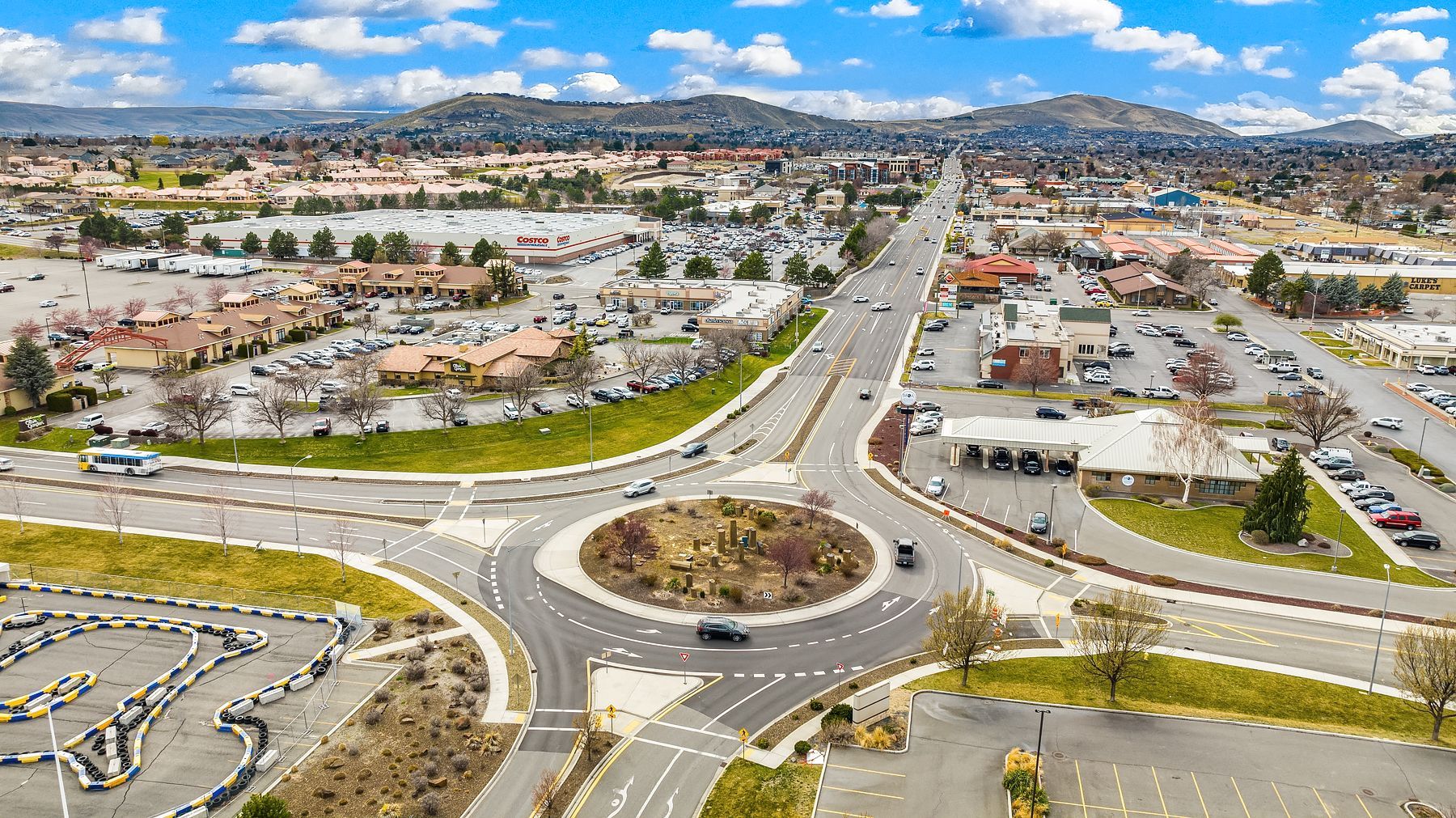 Aerial view of a large roundabout with multi-lane roads, cars, buildings, and mountains in the distance.