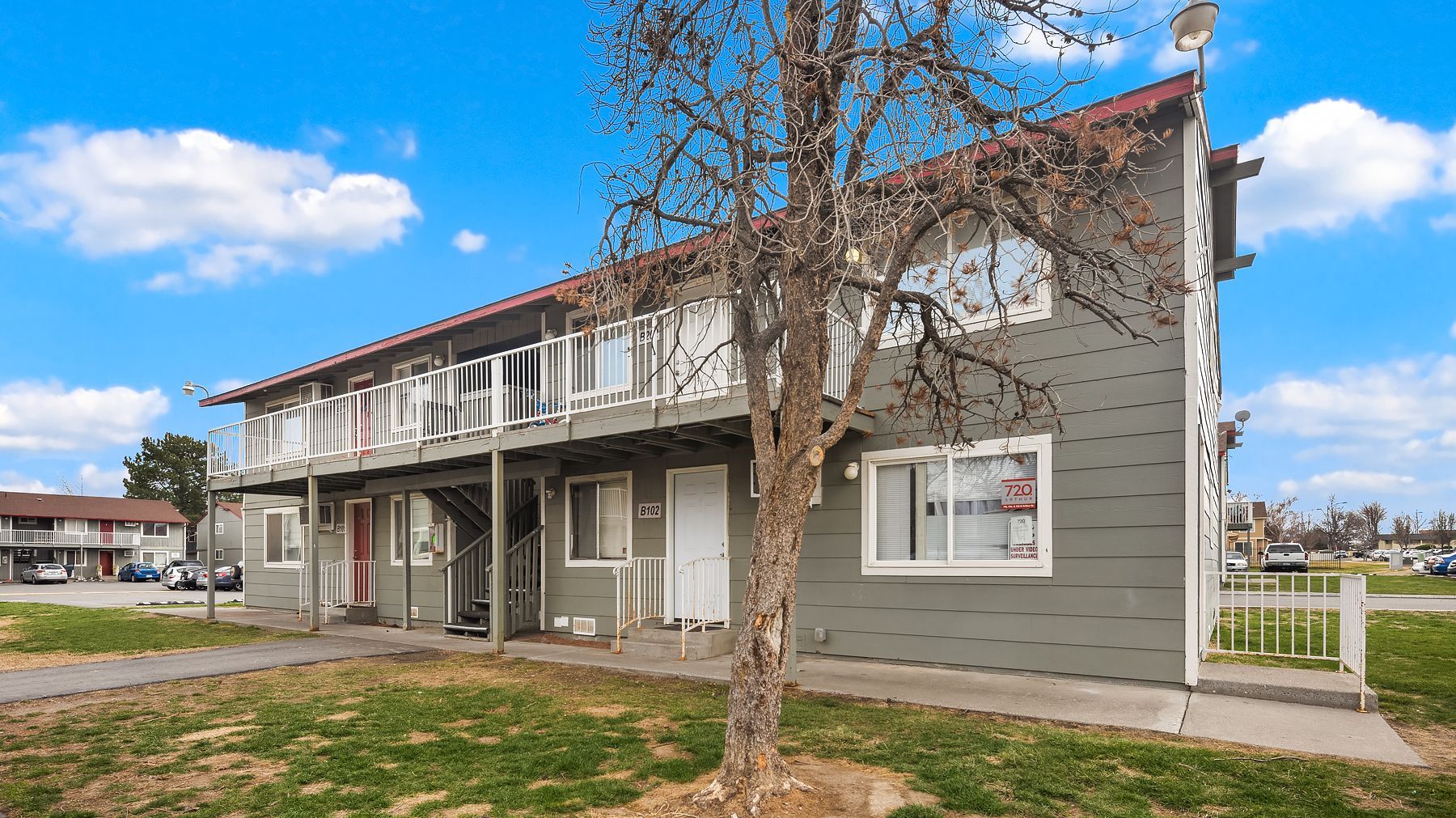 Two-story gray apartment building with white trim and a leafless tree in front under a blue sky