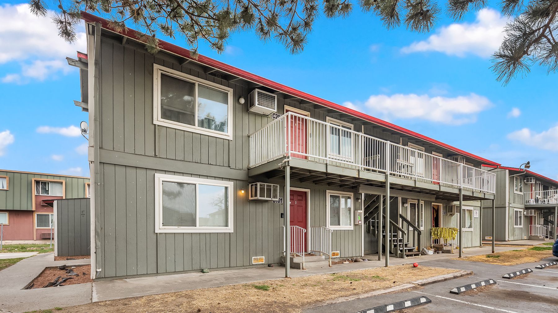 Two-story apartment building with gray siding, red trim, and exterior stairways under a blue sky