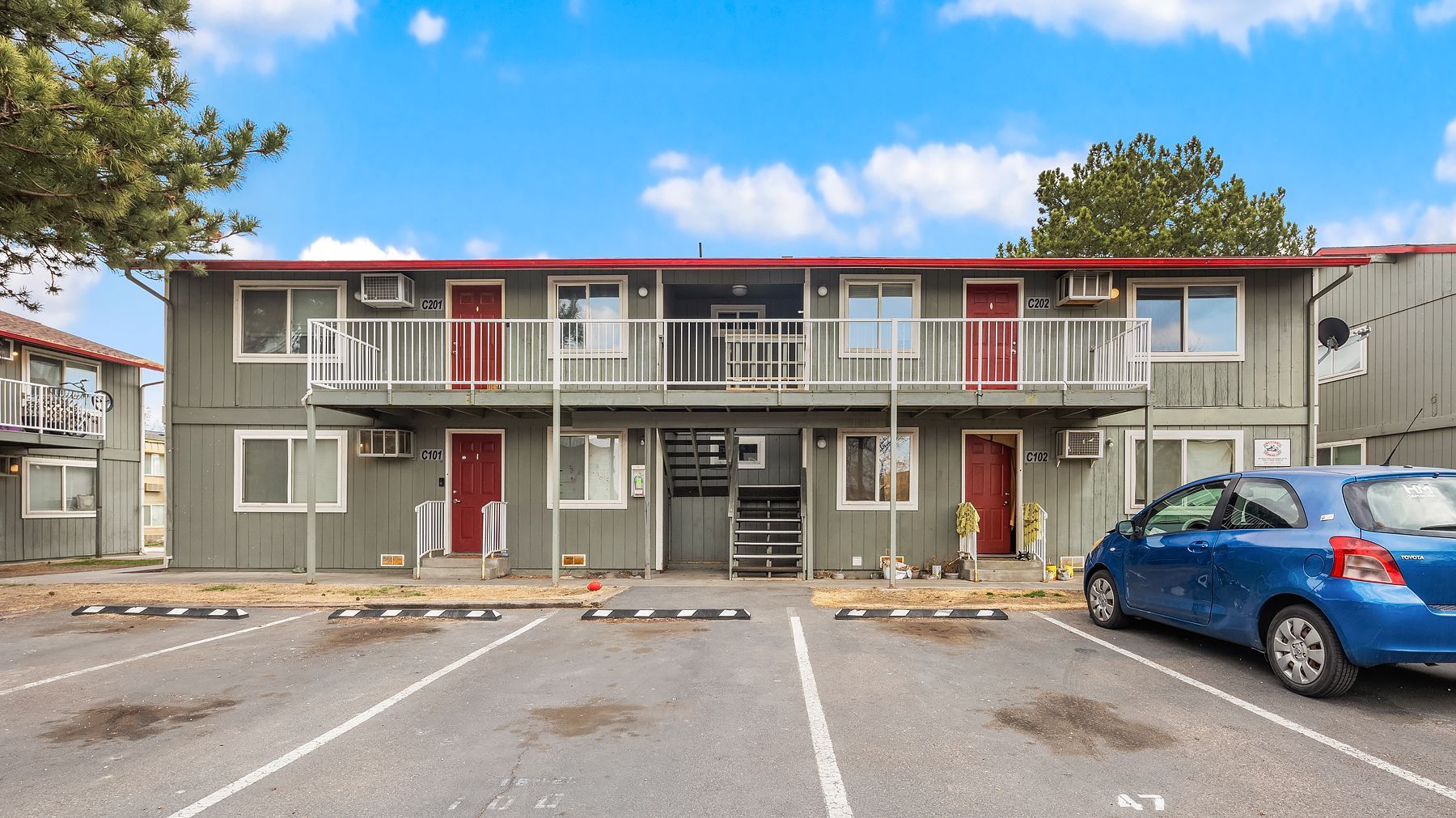 Two-story apartment building with balconies and parked cars in front under a blue sky