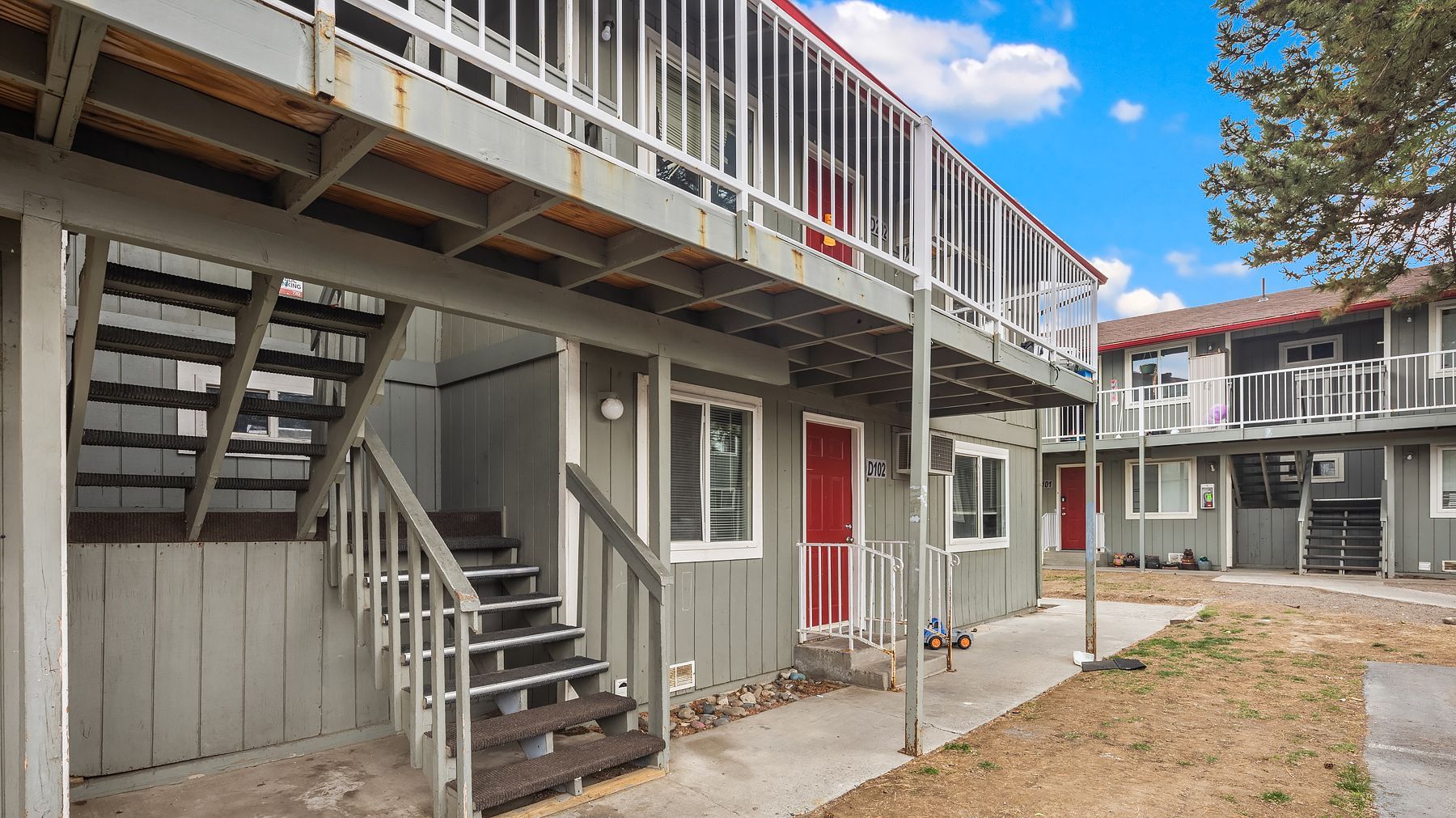 Two-story apartment courtyard with exterior staircases, white railings, and a red door under a balcony.