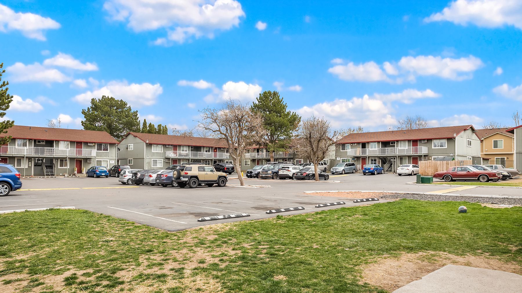 Apartment parking lot with cars, trees, and a grassy foreground under a partly cloudy blue sky
