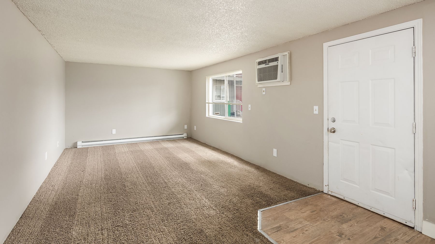 Empty beige living room with carpet, window, wall vent, and white front door