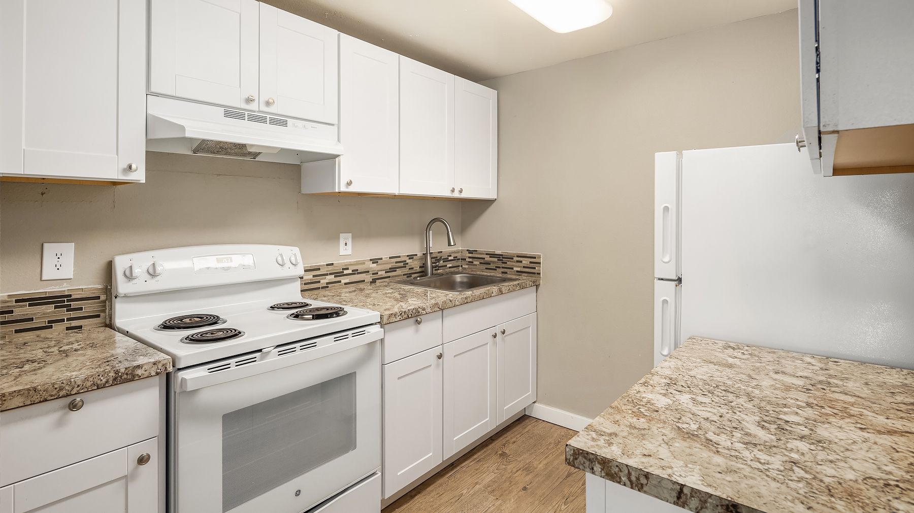 Empty white kitchen with granite countertops, white cabinets, stove, fridge, and wood flooring.