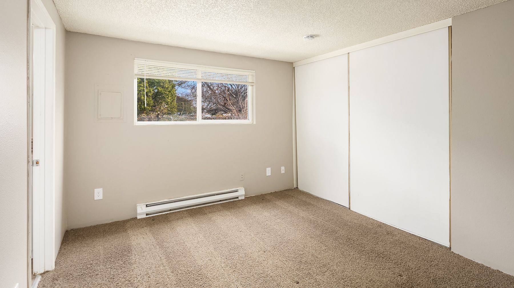 Empty beige bedroom with carpet, window, baseboard heater, and sliding closet doors