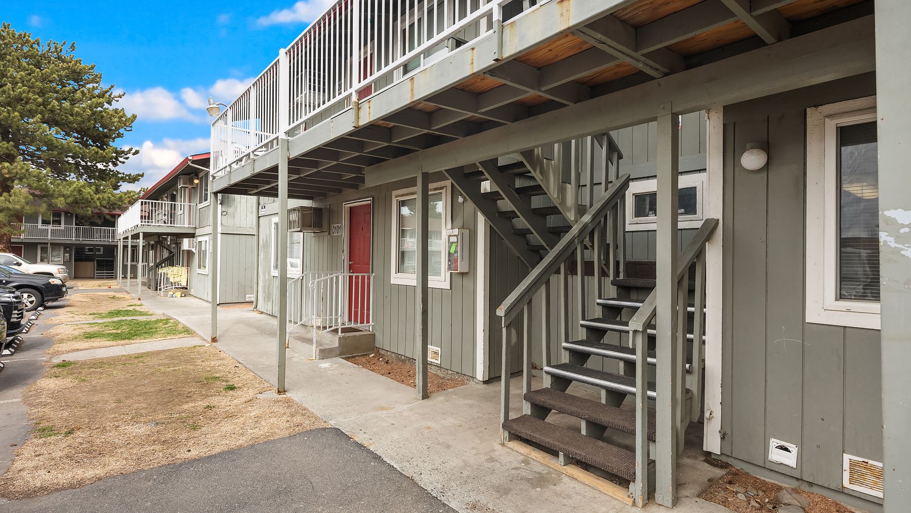Outdoor apartment walkway with stairs, railing, and doors along gray building under a blue sky