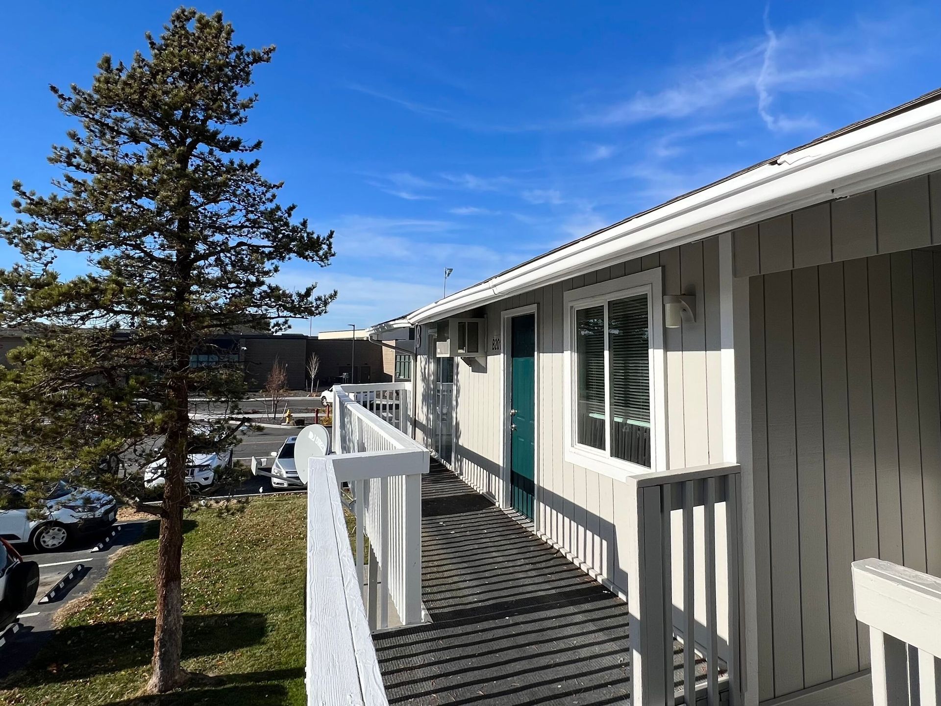Apartment balcony walkway with white railings overlooking a parking lot and trees under a blue sky