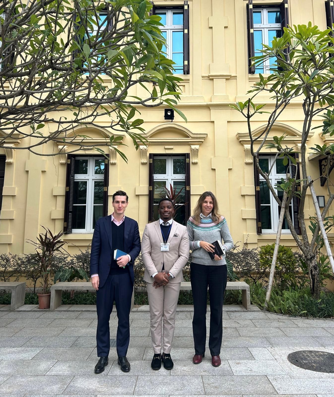 Drei Personen stehen vor einem gelben Gebäude mit Fenstern. Sie lächeln.