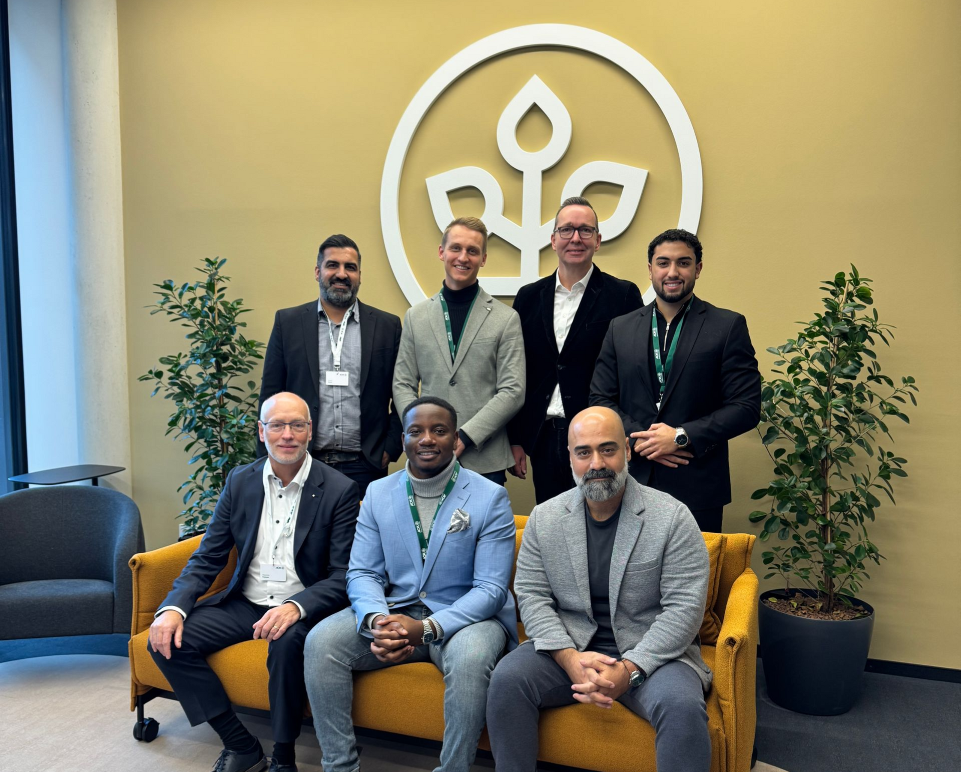 Eine Gruppe von Personen posiert vor einem Firmenlogo. Sie tragen Anzüge und lächeln in einer Büroumgebung.