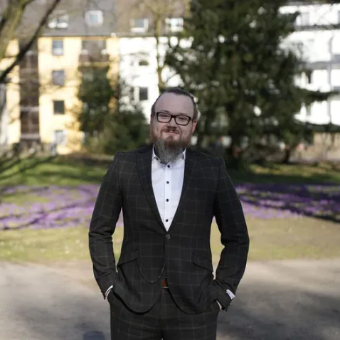 Mann im karierten Anzug, Hände in den Taschen, lächelt im Freien zwischen Blumen und Gebäuden.