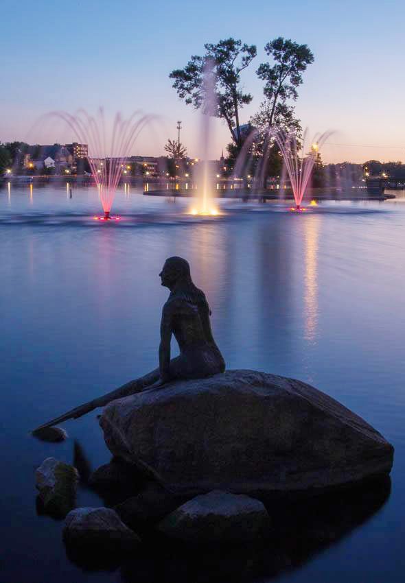 A statue of a mermaid is sitting on a rock in front of a fountain.