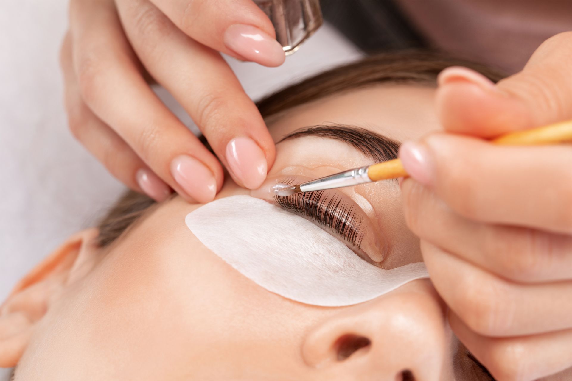 Close-up of a person getting a lash lift. An esthetician uses a brush and tool on the closed eyelid with protective pads.