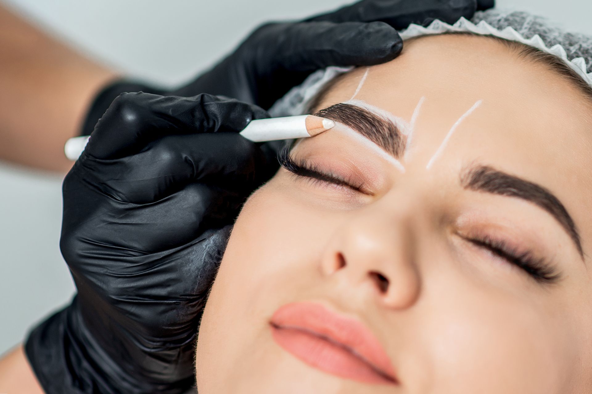 Eyebrow shaping: A person has eyebrows outlined with a pencil while another person, wearing black gloves, holds the pencil.