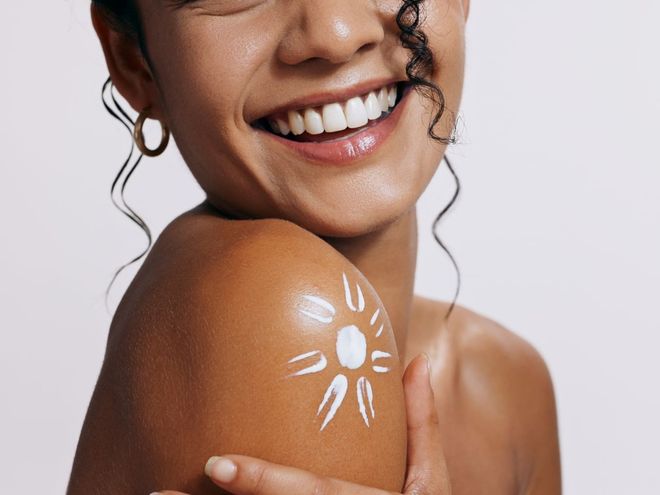 A woman is smiling and applying sunscreen to her shoulder.