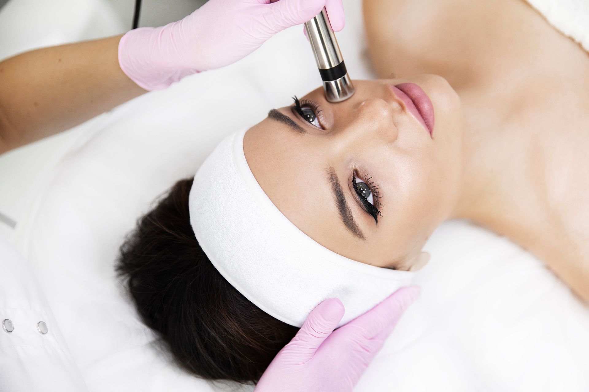 Woman receiving facial treatment; device on nose, pink gloves, white headband.