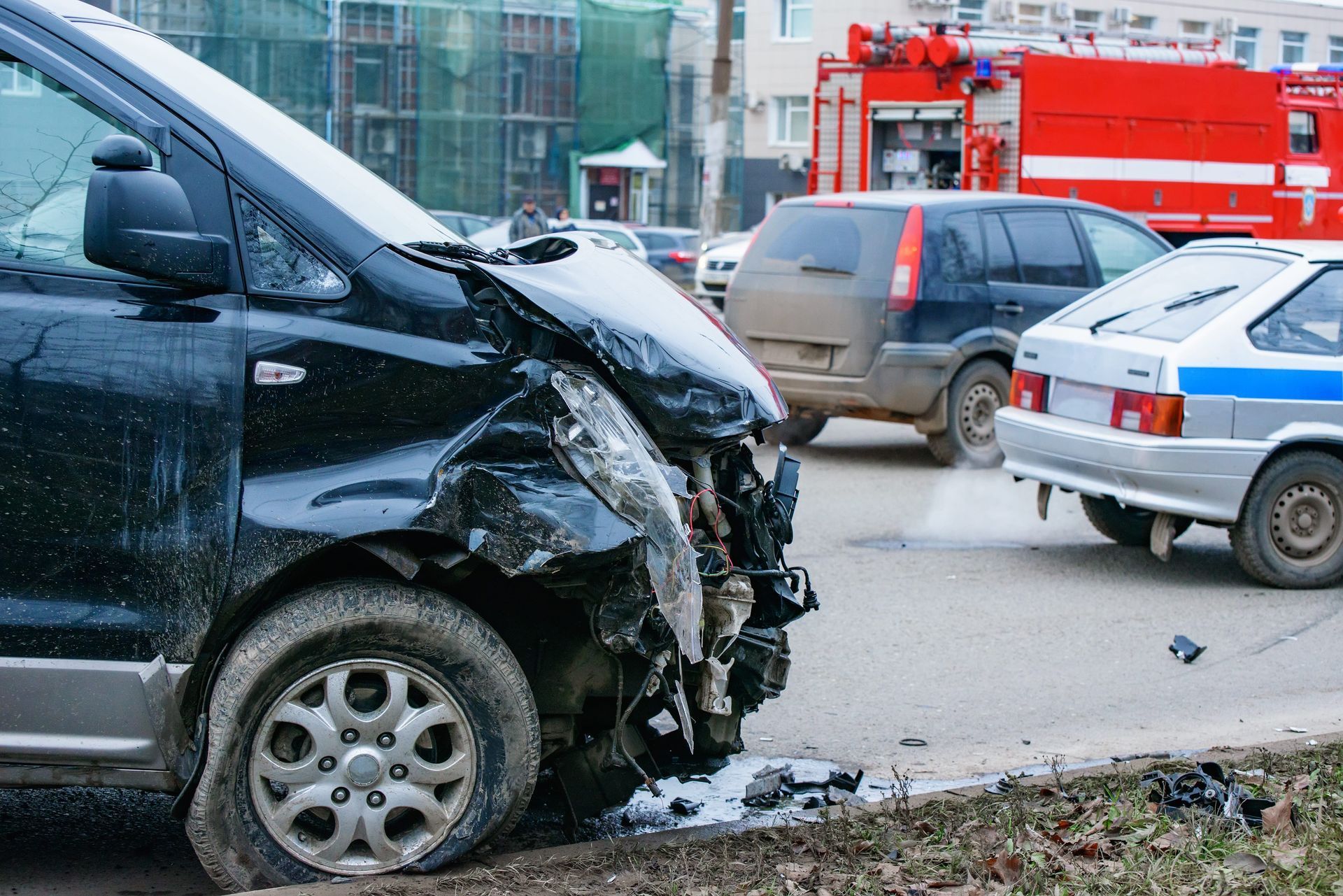 Damaged black van after a crash, with a red emergency vehicle and parked cars in the background