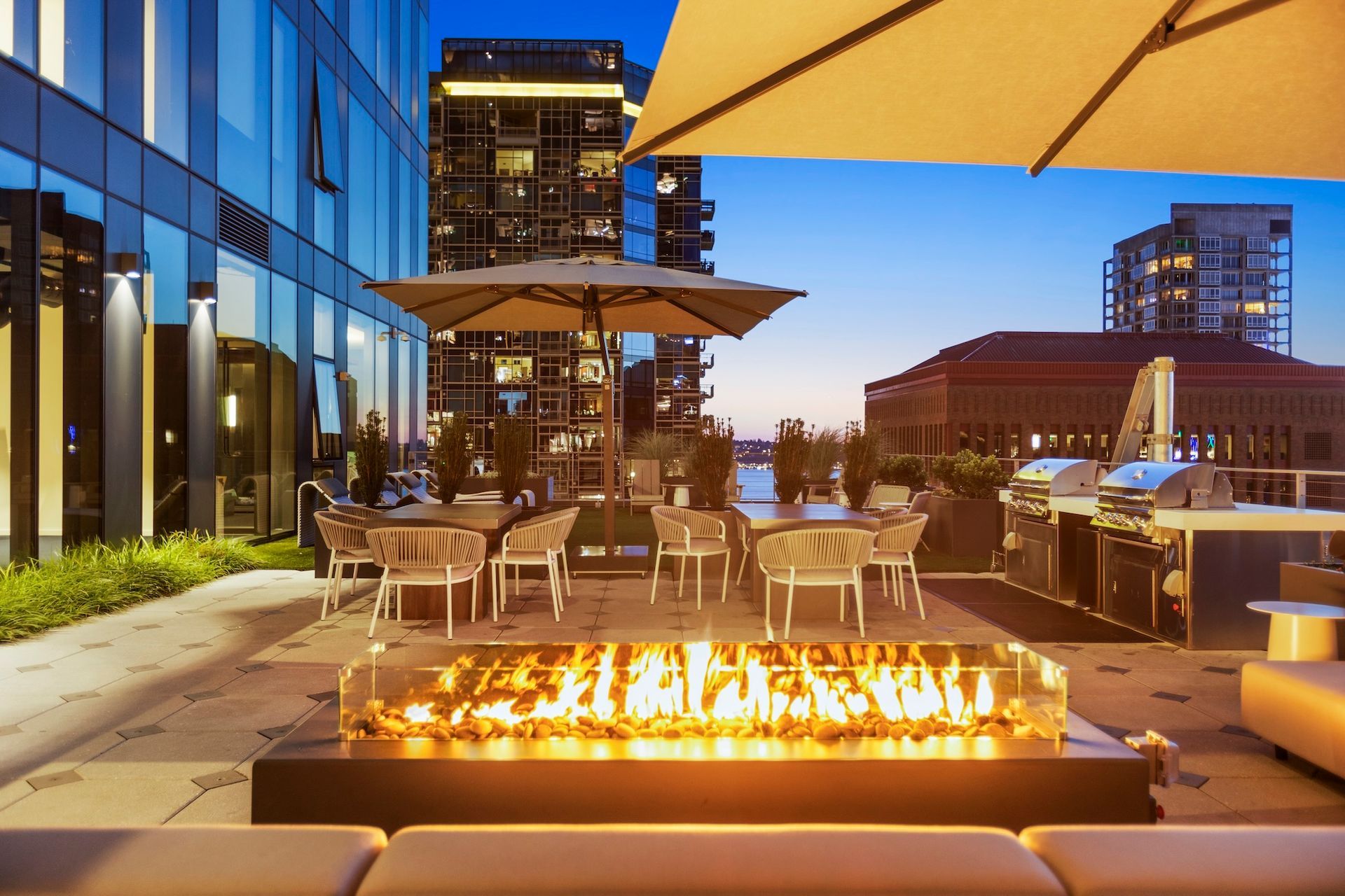 Rooftop patio at dusk with fire pit, outdoor dining tables, umbrellas, and city skyline in the background