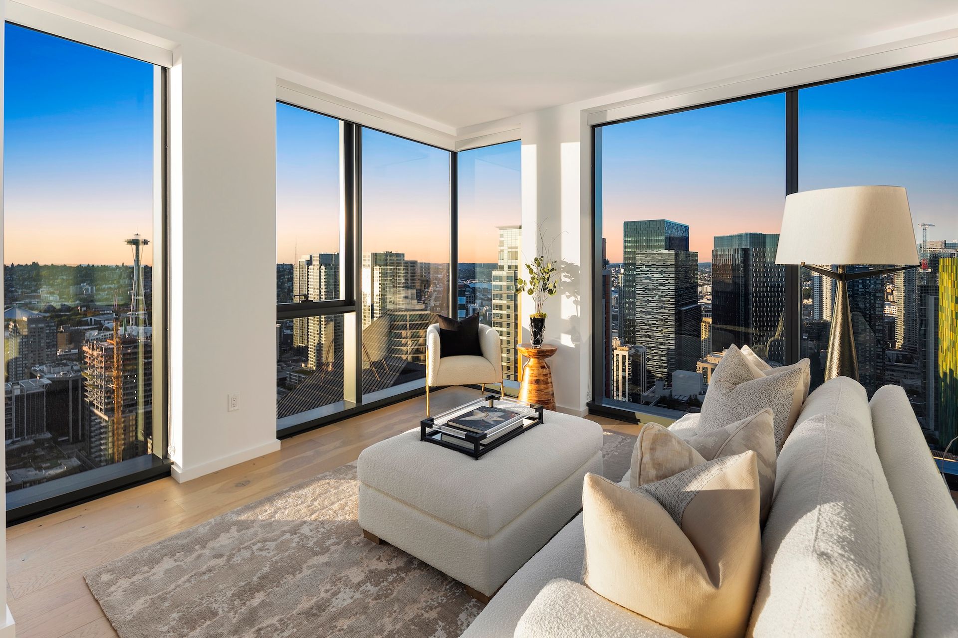 Modern living room with floor-to-ceiling windows, city skyline views, and neutral-toned sofa and ottoman