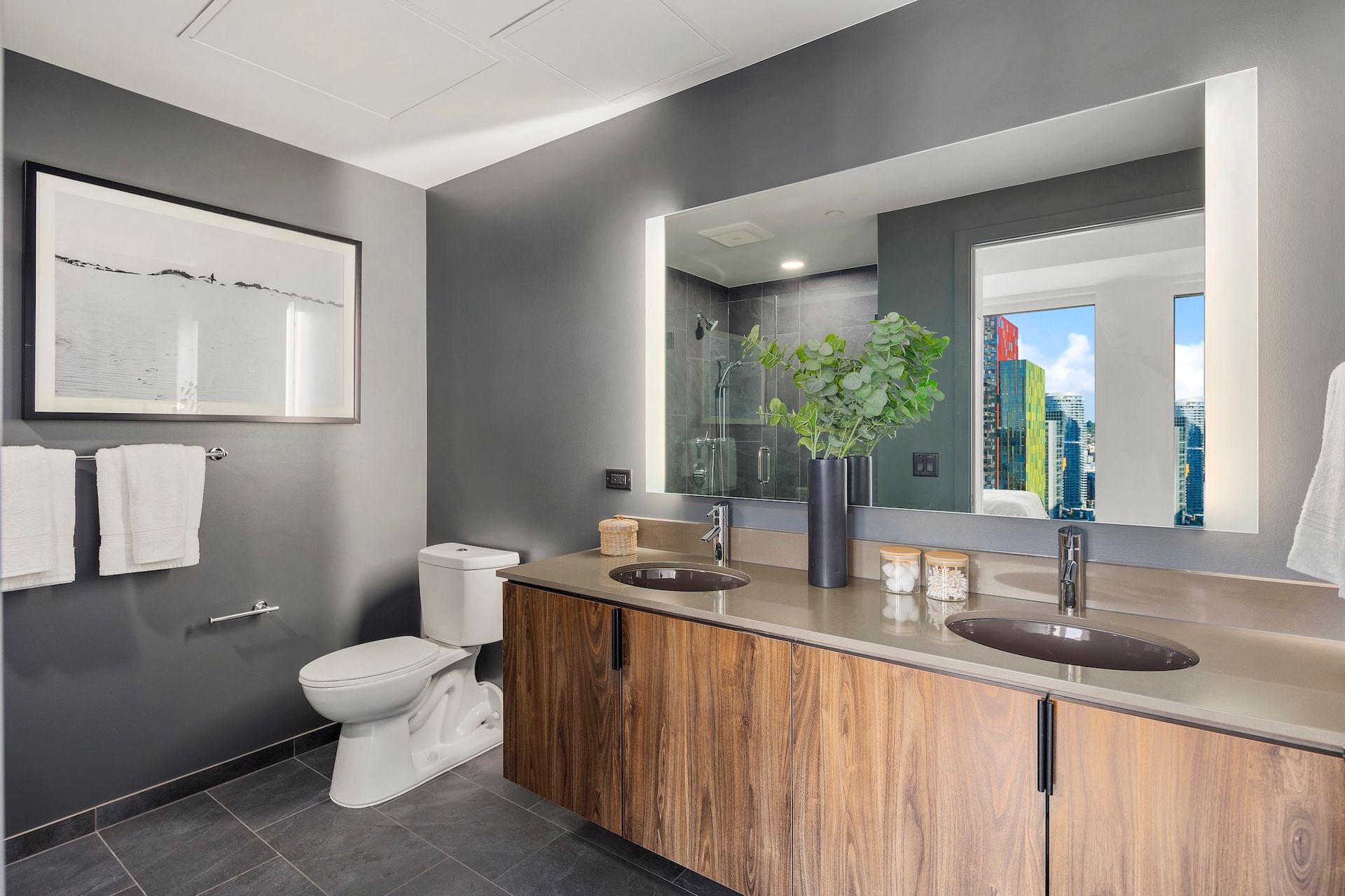 Modern bathroom with wood vanity, large mirror, toilet, and window view of greenery and buildings